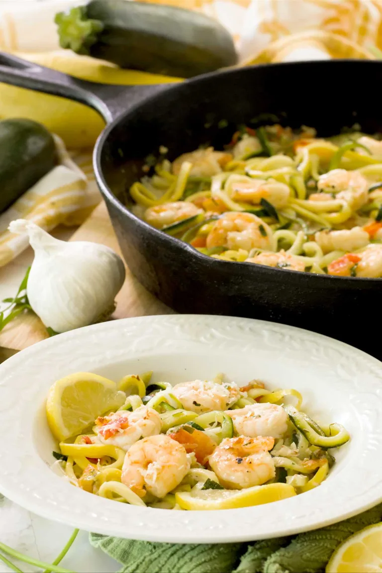 A white bowl of lemon garlic shrimp scampi with zucchini noodles in front of a cast-iron skillet.