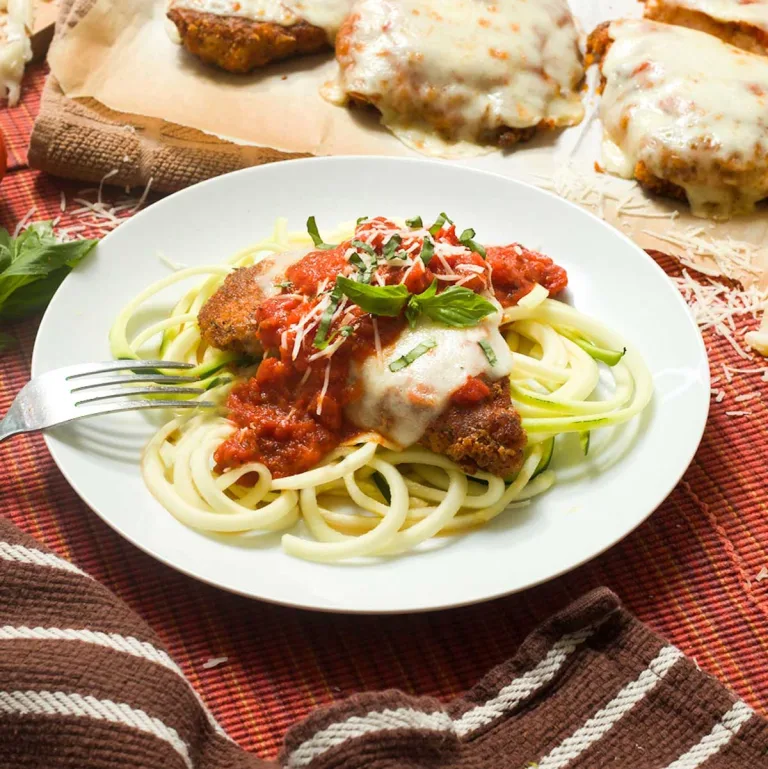 Gluten-free chicken parmesan with marinara sauce and mozzarella cheese on zucchini noodles on a white plate.