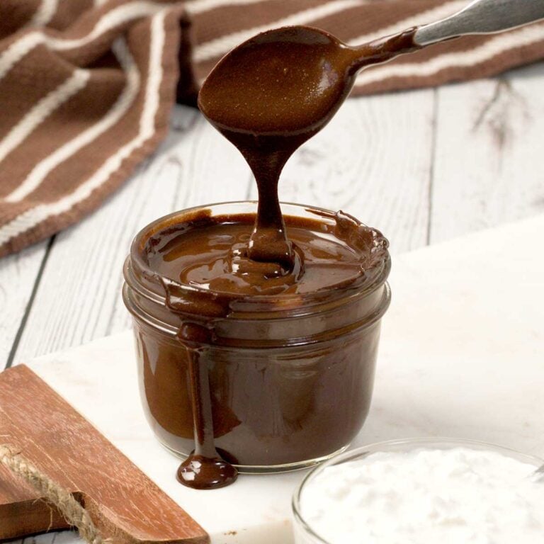 A close-up of a spoon dripping protein cottage cheese chocolate sauce in a mason jar on a white cutting board.