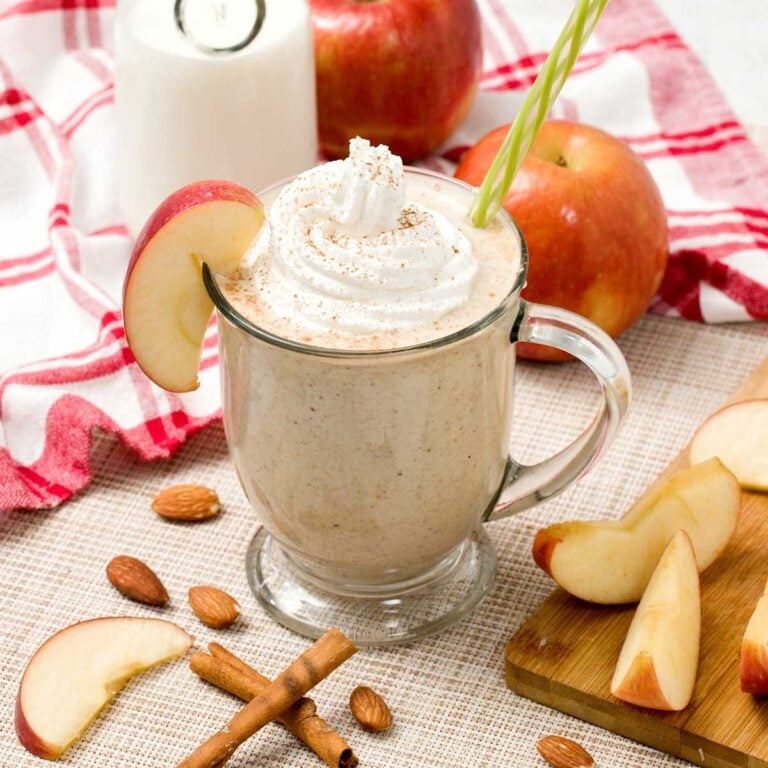 An apple pie smoothie in a glass with whipped cream on top with a green straw and slice of apple.