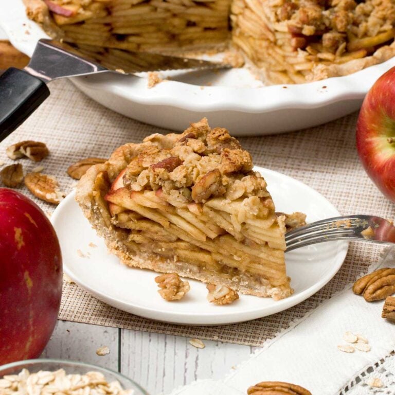 Slice of gluten-free apple crumble pie on a white plate next to an apple.