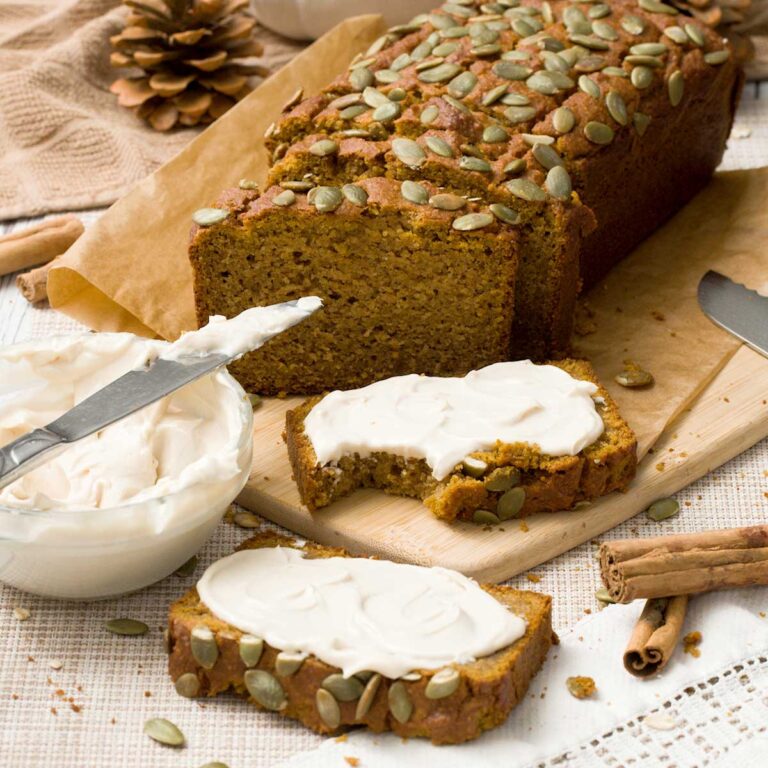 Two slices of gluten-free pumpkin bread with cream cheese frosting spread on them in a bite taking out of one next to a sliced loaf of bread.