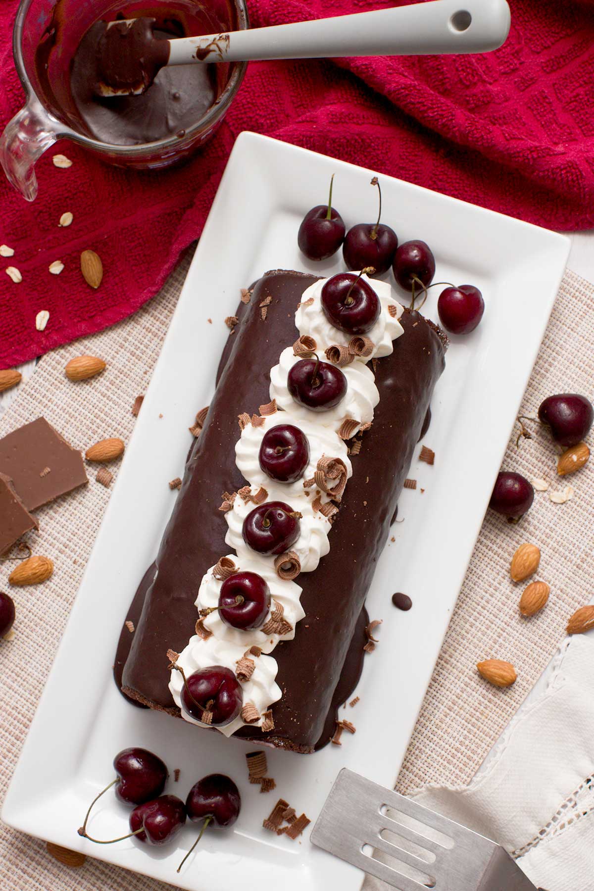 A gluten-free chocolate Swiss roll with cherries and whipped cream on top on a cake pan.