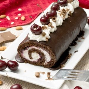 A gluten-free chocolate Swiss roll with cherries and whipped cream on top on a cake pan with a spatula.