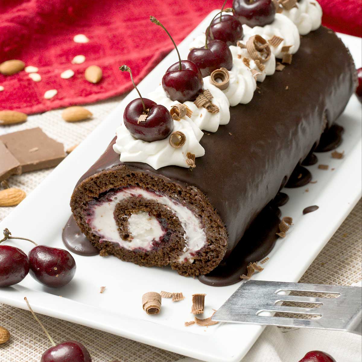 A gluten-free chocolate Swiss roll with cherries and whipped cream on top on a cake pan with a spatula.