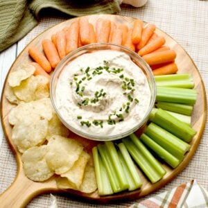 A bowl of french onion dip with greek yogurt surrounded by cut veggies and potato chips.
